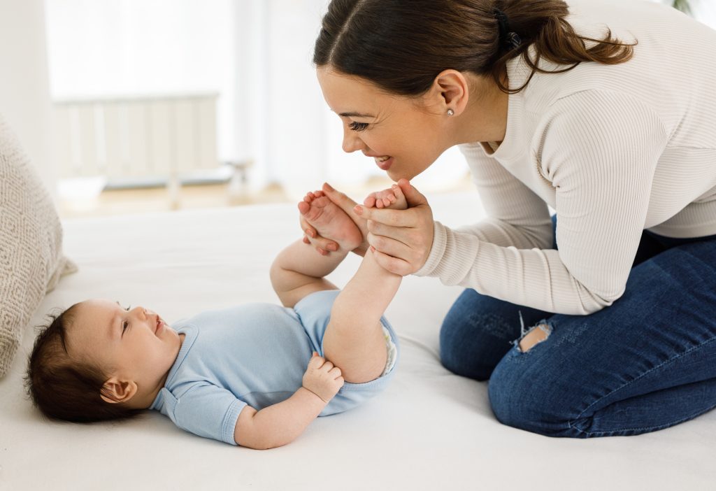 infant lying on back, mother holding infant's feet and smiling down at infant