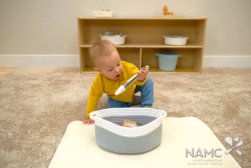 Infant in Montessori environment at a work mat with a basket of objects in front of him, examining tongs from the basket