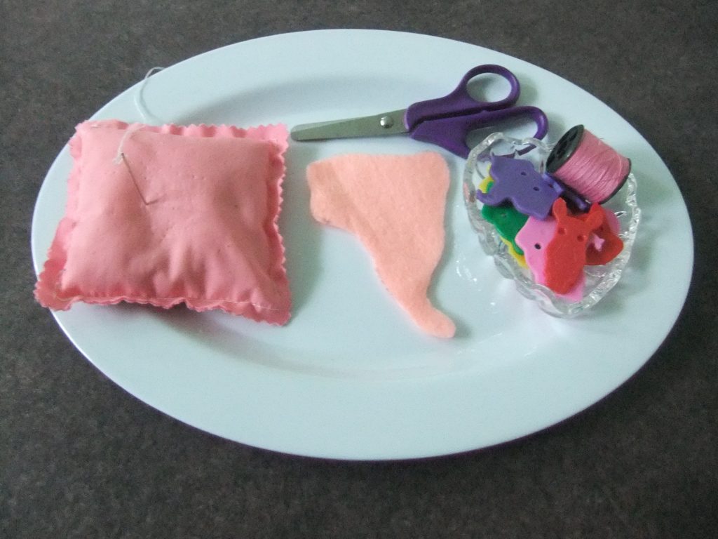 materials for South America sewing activity: pin cushion with threaded needle, child scissors, felt shape of South America, dish of animal shape buttons, pink thread
