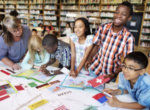 Elementary students studying geography. Maps, atlases, and flags scattered on table