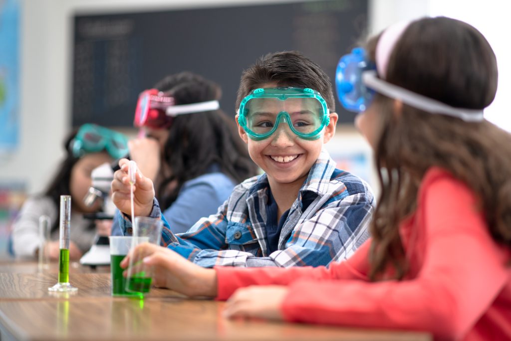 Two elementary students conducting a science experiment. They are wearing safety goggles and sitting at table with green liquid in front of them