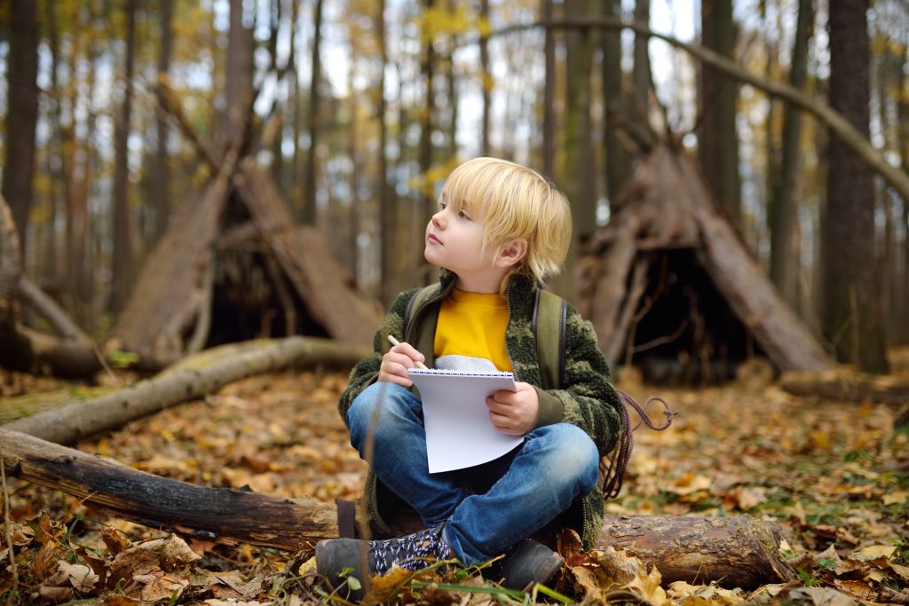 young boy sitting on forest floor, writing and looking thoughtful