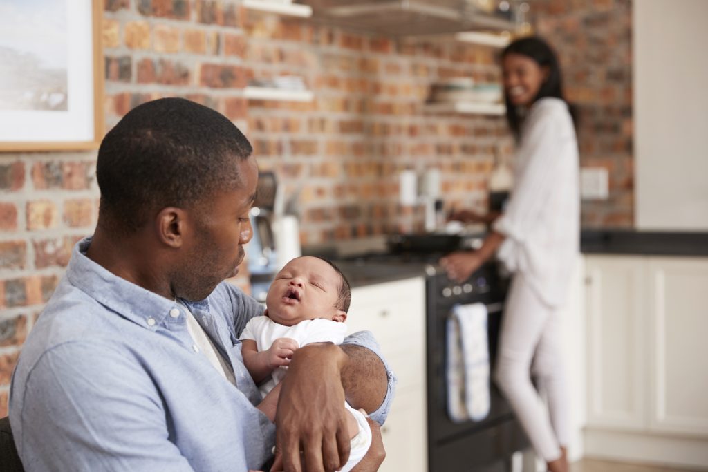 father sitting and  holding newborn, mother standing in  kitchen, smiling at them