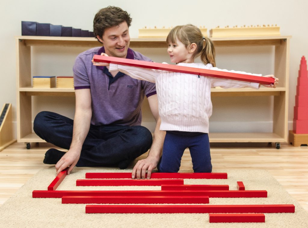 Montessori teacher and child sitting at a work mat with the Red Rods. Child is holding one of the largest rods in outstretched arms.