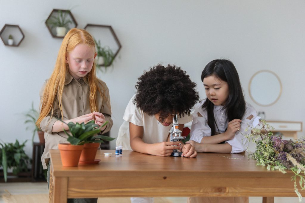 Three elementary students at table. Once is looking through a microscope. A variety of plant pots are also on the table