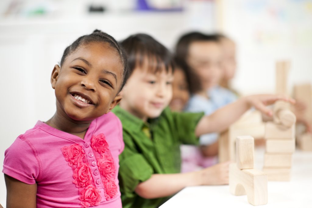 Montessori children sitting at table with building blocks. One student smiling at camera