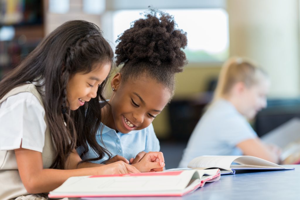 Two students sitting at a table and reading a book together, smiling