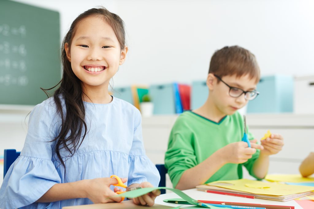 elementary age girl and boy working at a table in the Montessori classroom. Girl is smiling at camera