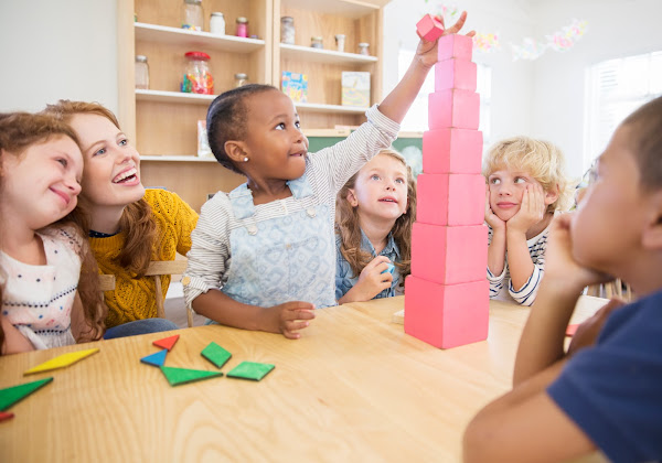 Group of Montessori Lower Elementary students and teacher sit around table. One girl places a cube on top of Pink Tower.