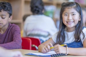 Elementary student smiling at camera, sitting at desk, preparing to write in her journal